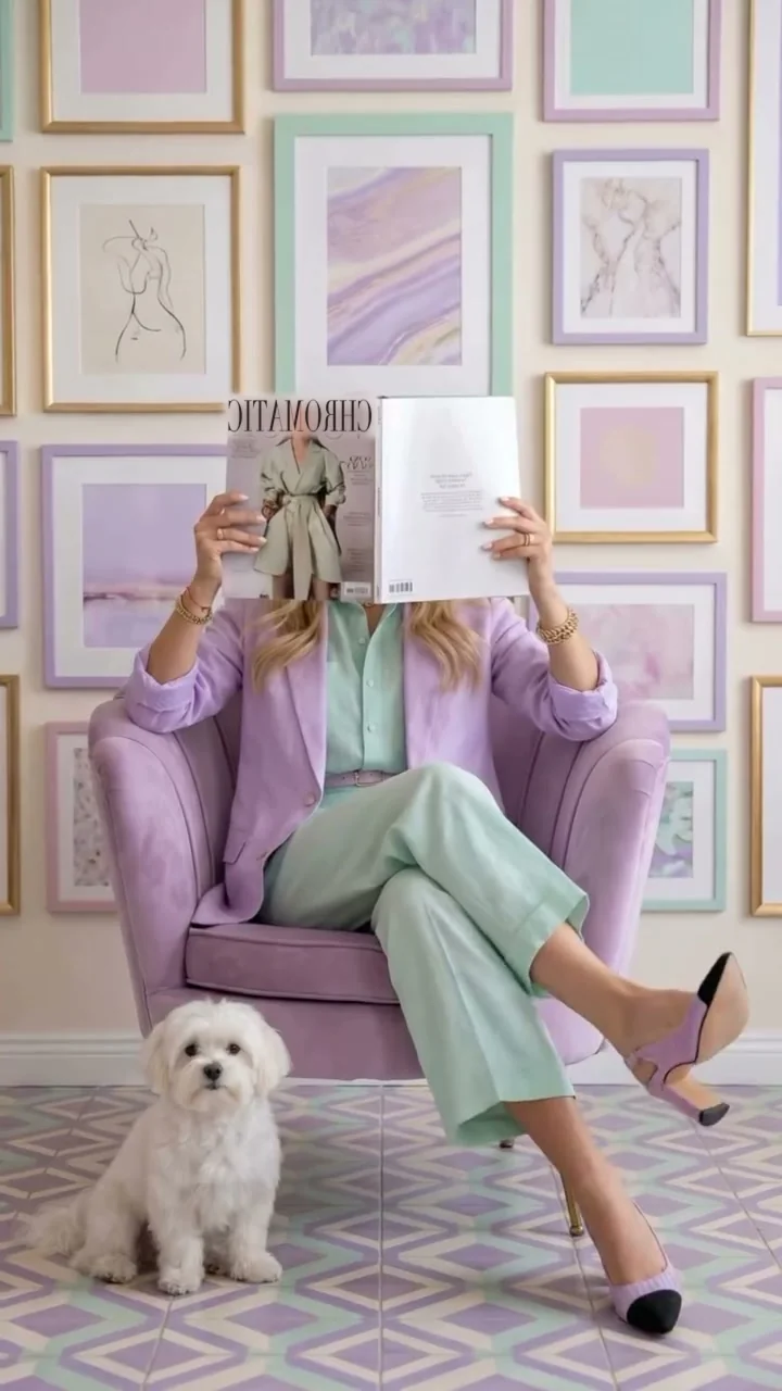 A woman in a lilac jacket and mint trousers sits in a lilac velvet armchair, reading an open magazine whose right-hand page is blank; a white Maltese sits on a lavender rug.
