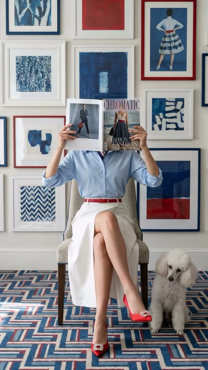 A woman in a sky-blue shirt, ivory skirt, red belt and red heels sits on a cane chair before cobalt cut-paper prints; a white standard poodle sits beside her on a blue zigzag rug.
