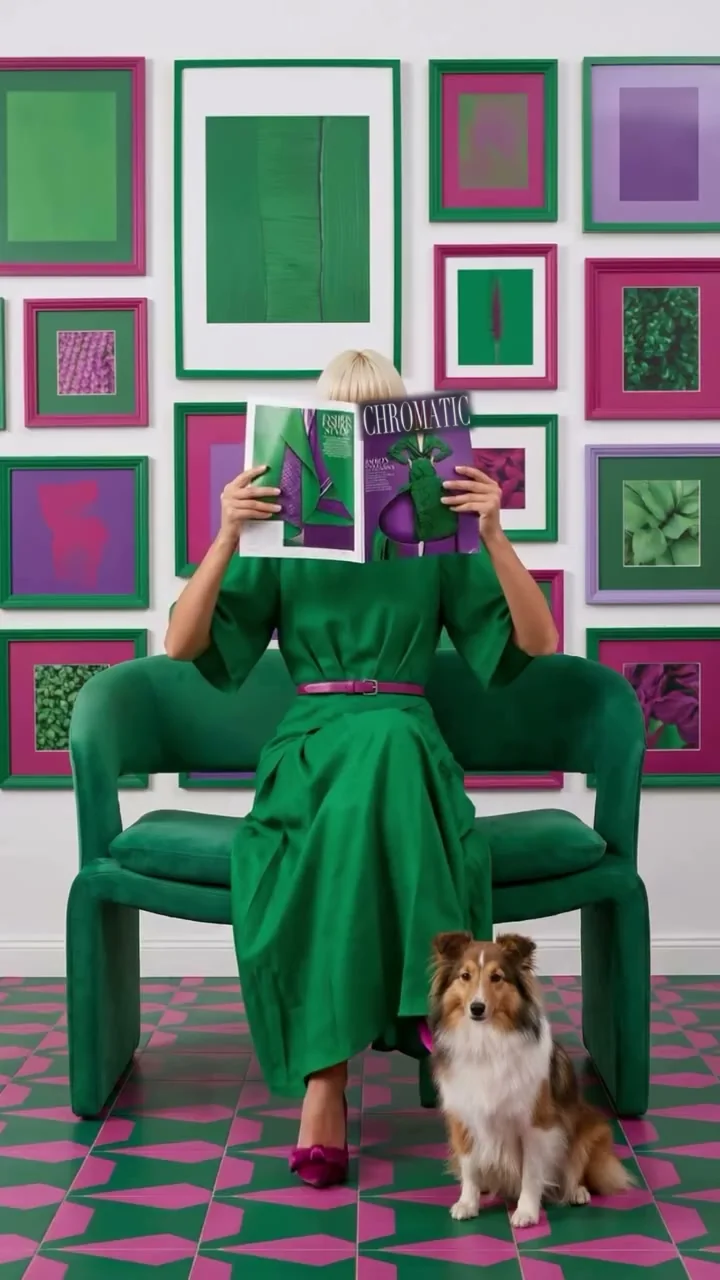 A woman in an emerald crêpe dress with a magenta belt sits on a green bouclé loveseat, framed by green and magenta prints; a Shetland sheepdog sits on a magenta and green tiled floor.