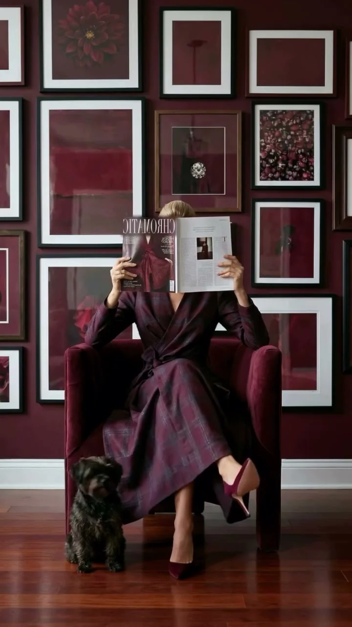 A woman in a plum and claret check wrap coat sits in an oxblood velvet armchair against dark claret walls hung with specimen frames, reading an open magazine; a small black dog sits at her feet.